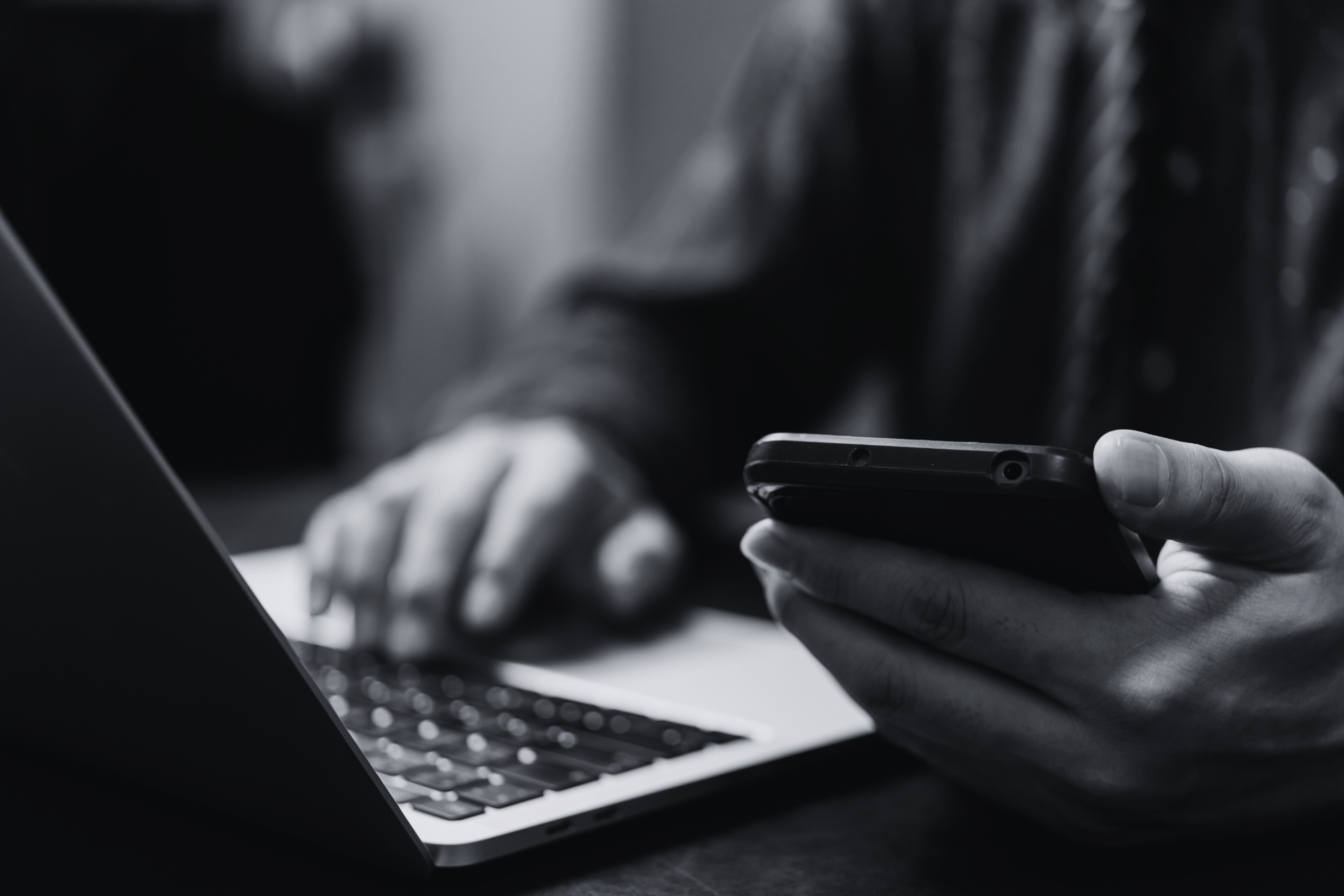 Schwarz-weiß-Foto einer Person, die am Laptop tippt und parallel ein Smartphone für Cyber-Security-Kontakt nutzt.